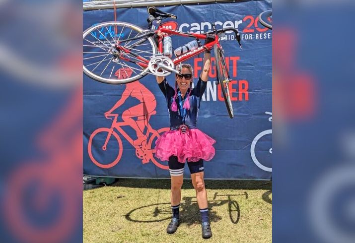 Former Peel Health Campus breast cancer patient Brooke Glatz wears cycling clothes and a bright pink tutu. Above her head she holds a racing bicycle.